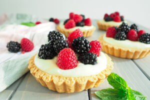 Tartelettes mit Beeren (Himbeeren und Brombeeren) angerichtet auf grauem Holzuntergrund. Ein Tartelette im Vordergrund und drei im Hintergrund. 