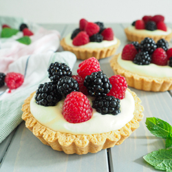 Tartelettes mit Beeren (Himbeeren und Brombeeren) angerichtet auf grauem Holzuntergrund. Ein Tartelette im Vordergrund und drei im Hintergrund. 