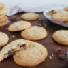 Cookies mit Schokostückchen auf braunem Holzuntergrund. Schokolade im Vordergrund. Teller mit Keksen und Stapel mit Keksen im Hintergrund.