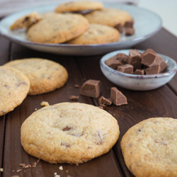 Cookies mit Schokostückchen auf braunem Holzuntergrund. Schokolade im Vordergrund. Teller mit Keksen und Stapel mit Keksen im Hintergrund.