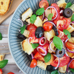 Tomaten-Brot-Salat mit Oliven, Zwiebel und Basilikum angerichtet in grau gestreifter Schale. Drum herum Tomaten, Basilikum und geröstetes Baguette auf rustikalem Holzuntergrund.